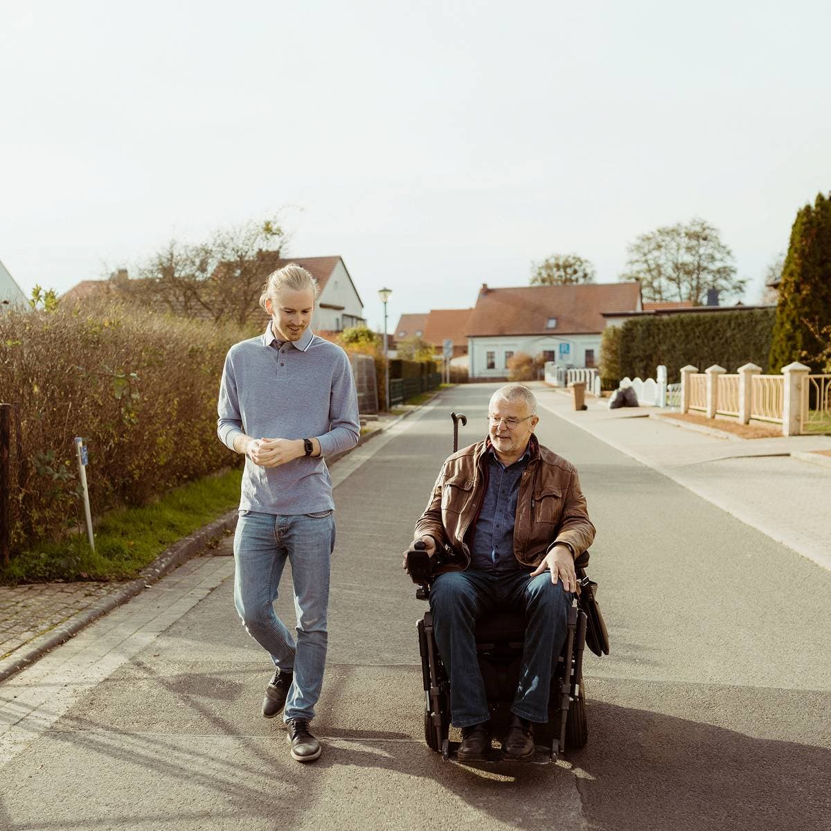En man i elrullstol och en gående kille pratar medan de tar sig fram på gatan i ett villaområde.