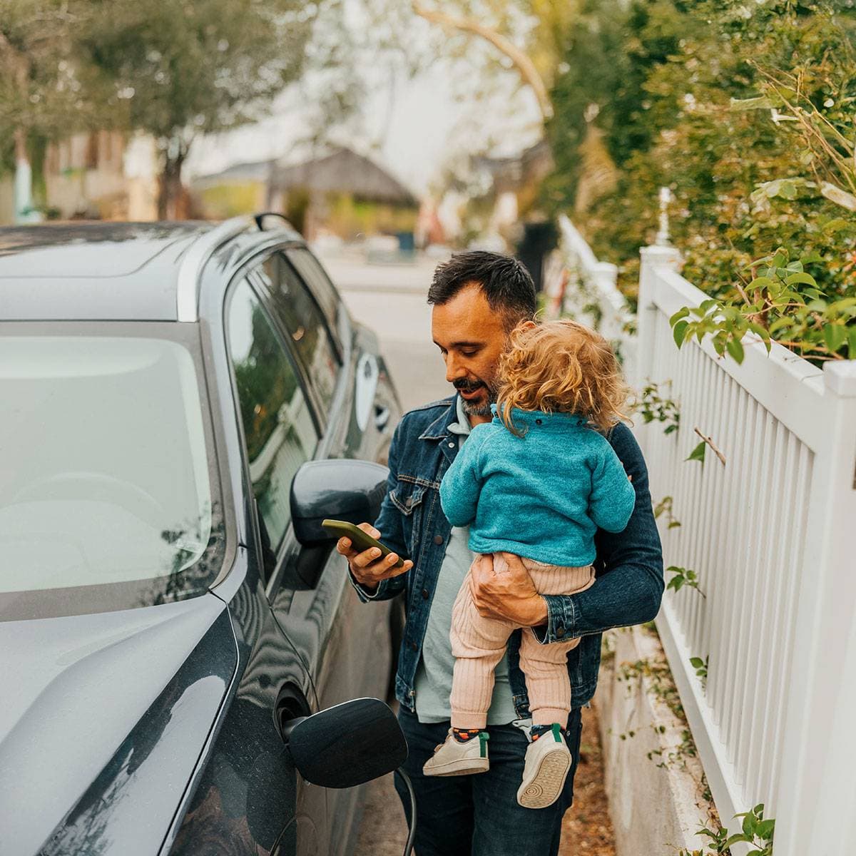 En man som står intill en bil och har ett barn på ena armen och mobilen i den andra handen.