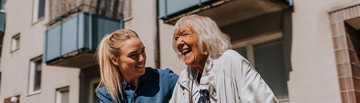 En äldre person använder en rullator utomhus medan en yngre person i blå tröja går bredvid och stöttar, båda framför ett bostadshus med balkonger i bakgrunden.