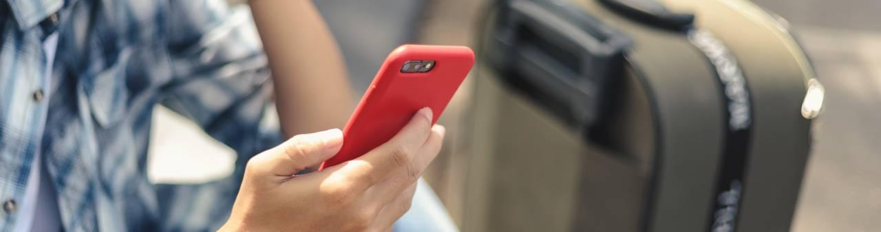 Person tittar i mobiltelefon, med en reseväska jämte sig.