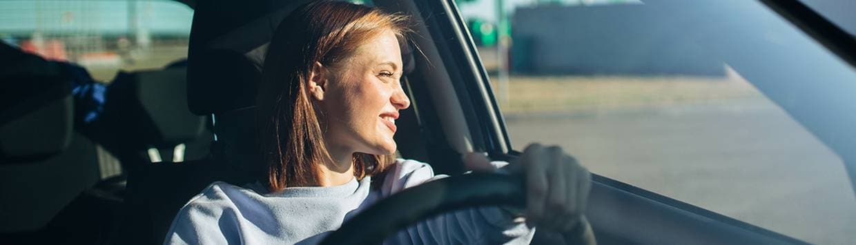 En leende tjej sitter i en bil, håller i ratten och tittar ut genom fönstret
