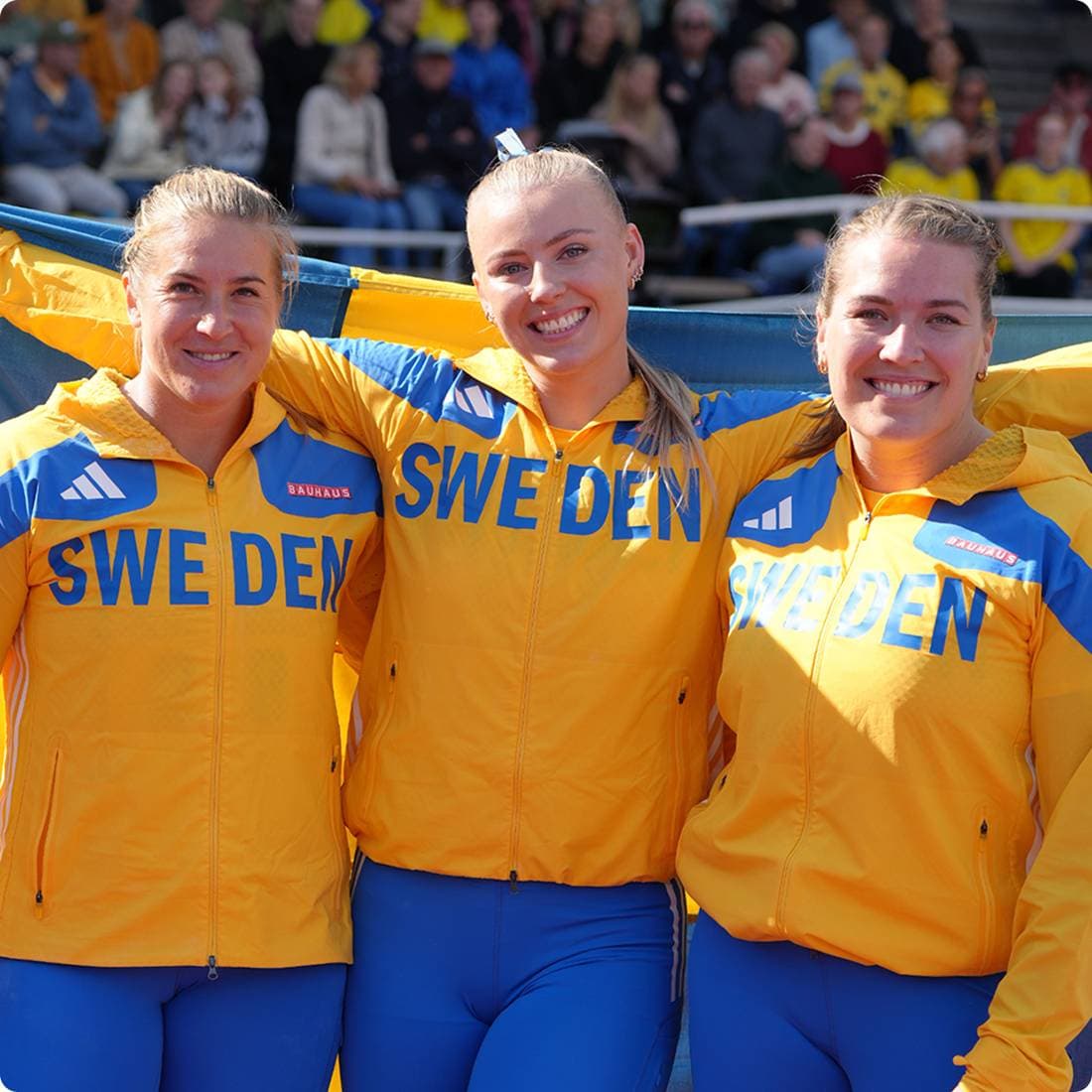 Tre glada kvinnliga friidrottare står tillsammans i sina landslagsdräkter och en håller i en svensk flagga bakom ryggen