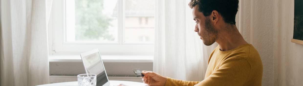 En man sitter framför sin laptop med ett kontokort i handen.