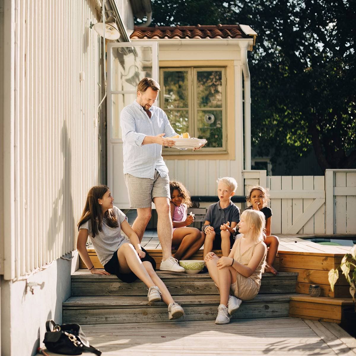 Barn sitter på en trappa utomhus, samt en man som går på trappan med ett stort fat.