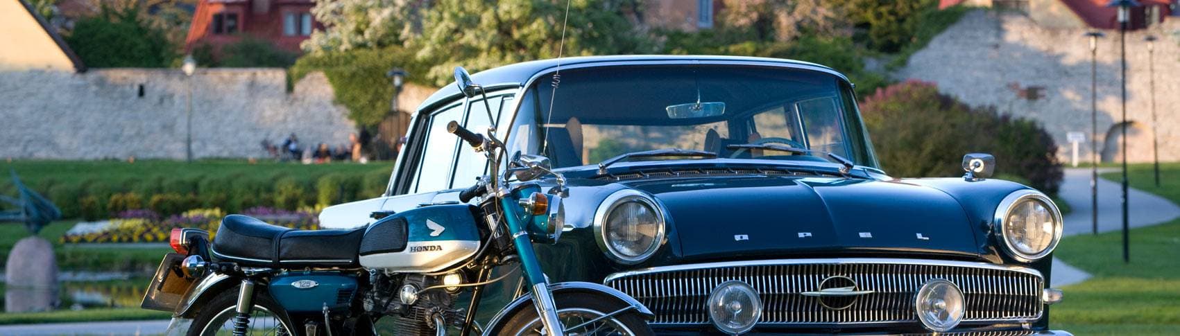 En äldre blå motorcykel av märket Honda och en äldre svart bil av märket Opel står parkerade i en park framför en mur.