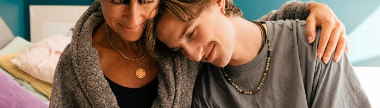 En kvinna med ett guldhalsband sitter bredvid sin tonårsson och håller armen om honom.