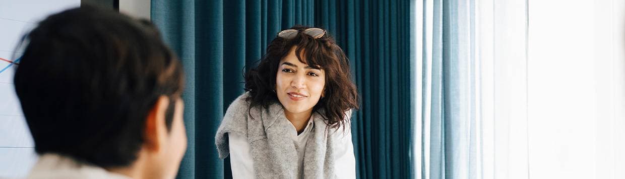 En kvinna med mörkt lockigt hår och glasögon på huvudet samtalar med en person i förgrunden, framför ett fönster med blå gardiner.