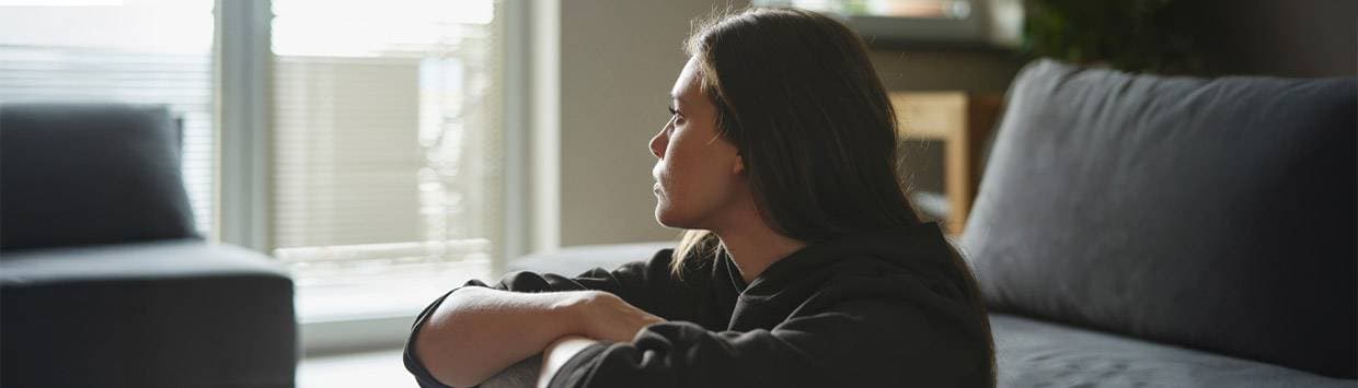 Kvinna sitter på golvet framför en soffa och tittar mot ett fönster där persiennerna skymmer utsikten.