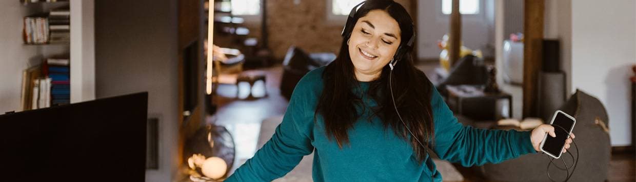 Kvinna som dansar i vardagsrum bredvid en TV-skärm och med en mobiltelefon i handen.