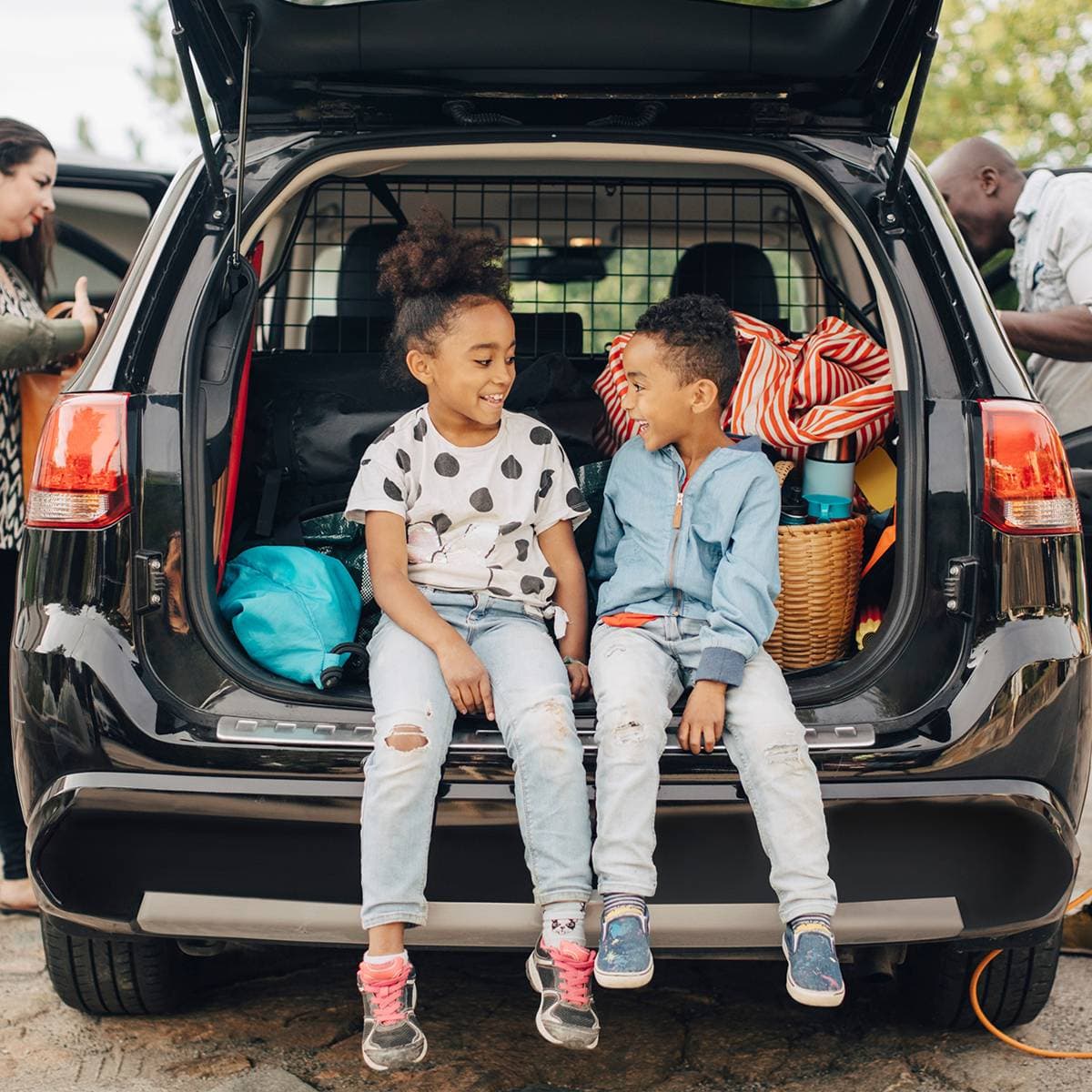 Två skrattande barn sitter i bakluckan på en bil och en man och en kvinna står på vardera sida utanför bilen vid framsätet