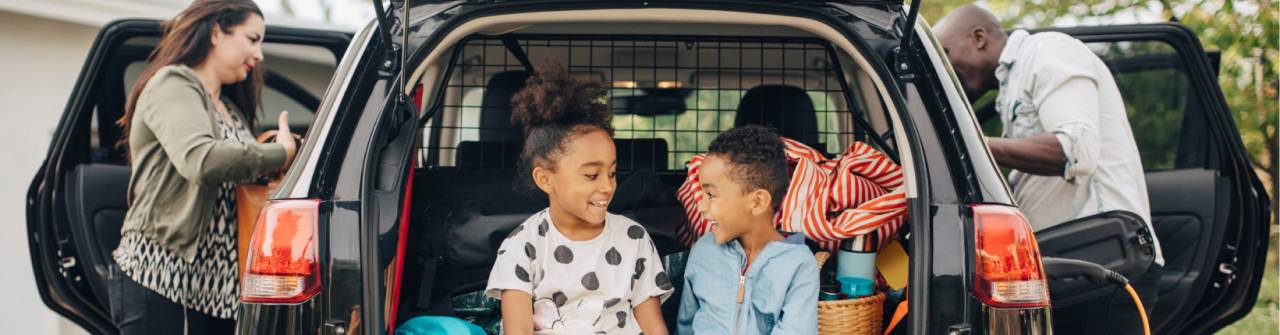 två barn som sitter i en baklucka medan föräldrarna packar bilen